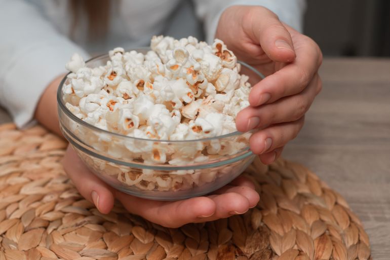 Woman Holding Bowl of Fresh Popcorn. Close-up of a woman's hands holding a transparent bowl filled with freshly popped popcorn, on a natural woven mat